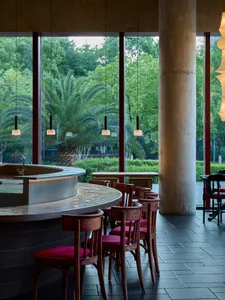 A modern restaurant with a bar, wooden chairs with red cushions, and a large window offering a view of trees outside