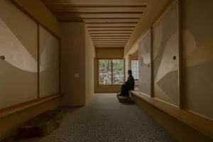 A woman sits on a stone bench in a calm and inviting room with a wooden floor and natural light from a window
