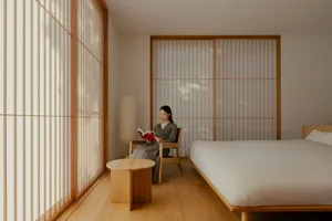 A woman sits in a cozy bedroom reading a book, surrounded by a bed and a chair near a window