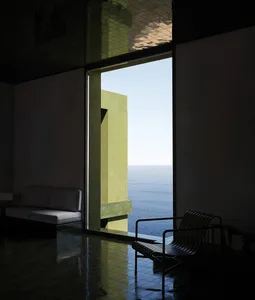 A well-lit living room with a couch and chair in front of a large ocean-facing window, featuring a mix of wood and metal furniture and decor