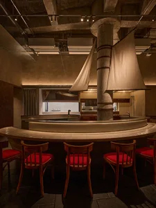 A modern restaurant with a curved bar and dining area, featuring a large dining table, wood and metal elements, and dim lighting creating a cozy atmosphere