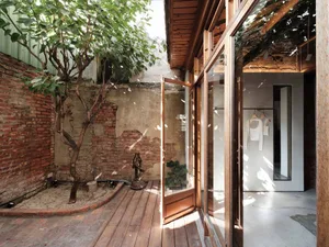 A modern living space with a large wooden door, glass window, and brick wall, featuring a tree in front and a dining table in the background, with bright and warm lighting