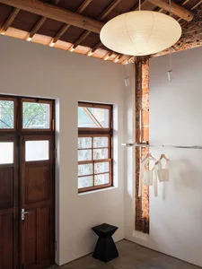 Minimal dressing area in an industrial loft featuring exposed red brick wall, large wooden windows, a black geometric stool, and a large white Akari paper lantern pendant hanging from a wooden beam ceiling.