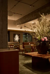 Interior view of a modern wabi-sabi style restaurant featuring wooden tables, upholstered chairs, exposed concrete elements, and a large floral centerpiece in a dark ceramic vase.
