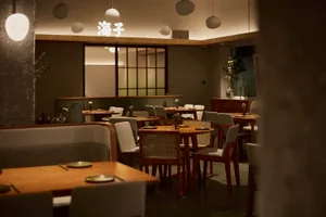 Interior view of a contemporary Japanese restaurant featuring wooden tables, upholstered and cane-back chairs, concrete pillars, and warm pendant lighting with Chinese characters illuminated on the back wall.