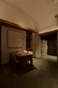 Modern wabi-sabi dining room featuring a wooden dining table with four upholstered chairs, a large topographic wood artwork on the wall, built-in wooden shelving, and a striking wall of hay bales under a high curved concrete ceiling with warm ambient lighting.