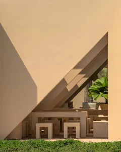 Modern brutalist outdoor lounge featuring raw concrete geometric furniture and stools under a slanted concrete canopy with tropical plants in the background.