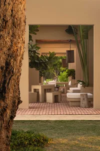 View from a garden into a minimalist outdoor living courtyard featuring geometric concrete tables, stools, and daybeds with white cushions on terracotta brick flooring, surrounded by lush tropical plants and hanging wooden lanterns.