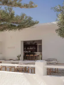 Outdoor dining area with a long travertine stone table and benches, wooden stools with woven seats, opening to a dark wood bar interior, framed by pine and olive trees under a clear blue sky.