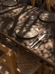 Close-up view of a weathered wooden outdoor dining table set with pewter plates, clear drinking glasses, and rustic cutlery on a red brick rest, with wooden chairs featuring woven seats under dappled sunlight and tree shadows.