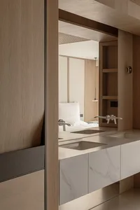 Modern minimalist bathroom with light oak cabinetry, white marble floating double vanity with two integrated sinks, and a large mirror reflecting a bedroom with white bedding.