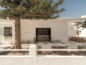 Outdoor dining courtyard with long stone table, wooden stools, whitewashed building, and a large pine tree under clear blue sky