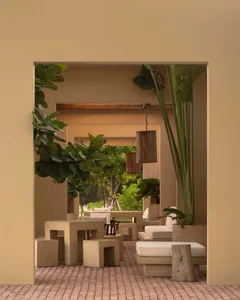View through a beige stucco archway into an outdoor courtyard patio with beige concrete modular sofas, square tables, hanging cylindrical wooden lanterns, large potted fiddle leaf fig and banana plants, on a terracotta brick floor.
