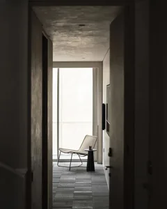 View through a doorway into a minimalist concrete hallway featuring a woven lounge chair, black side table, and large window with natural light.