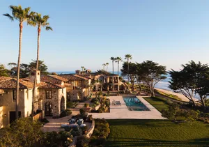 Aerial view of a luxurious Mediterranean-style stone villa with terracotta roof tiles, infinity swimming pool, manicured gardens, and tall palm trees overlooking the ocean at sunset.