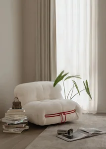 White boucle teddy-bear style lounge chair with red rope detail, positioned next to a stack of design books, a small handbag, an open magazine, and a potted plant in front of a bright window with sheer curtains.