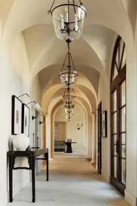 Long arched hallway in Tuscan style featuring multiple glass and iron lanterns, a distressed black console table with white ceramic vase, framed artwork, and limestone flooring leading to a seating area.