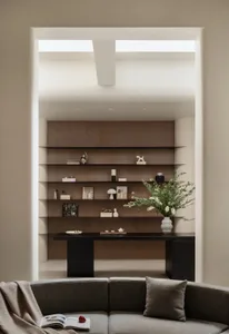 Modern minimalist living room featuring a curved grey sofa in the foreground, with a large built-in walnut wood shelving unit and a black console table in the background, decorated with books, sculptures, and a large white vase with green branches.