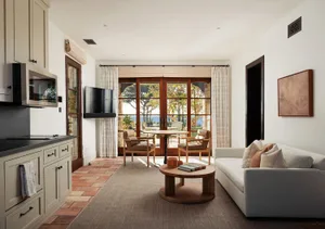 Bright open-plan living room featuring terracotta floor tiles, a light gray sofa, round oak coffee table, two leather armchairs, wall-mounted TV, and large glass doors opening to a coastal terrace with ocean view.