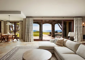 Bright coastal living room featuring a large L-shaped sectional sofa, round wooden coffee table, patterned area rug, and expansive ocean view through three brick arches leading to a patio.