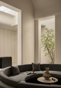 Modern minimalist living room featuring a large curved grey sectional sofa, round coffee table with sculptural vase, travertine walls, and a small tree in an open courtyard under a skylight.