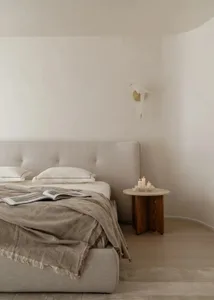 Minimalist bedroom featuring a large upholstered linen bed with beige throw blanket, wooden side table with candles, and a sculptural wall sconce against curved off-white walls.