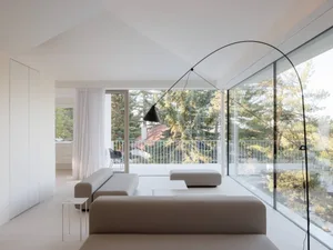 Modern minimalist living room featuring low-profile beige fabric sofas, a large black arched floor lamp, white walls, and floor-to-ceiling glass windows overlooking a lush green forest and balcony.