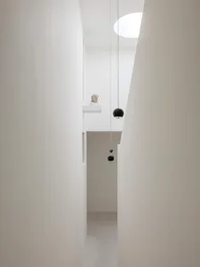 View looking up into a minimalist white double-height hallway featuring two black spherical pendant lights, a classical bust on a high ledge, and a large circular skylight.
