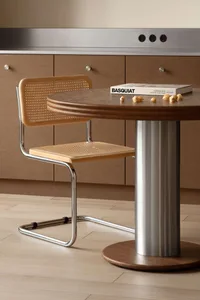 A light wood cane chair with chrome frame sits beside a round walnut pedestal dining table featuring a stainless steel column base, with a Basquiat book and walnuts on the tabletop.