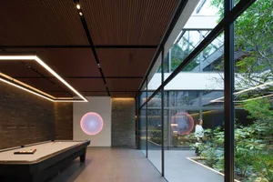 Modern indoor billiard room featuring a black pool table, linear LED lighting, dark wood slat ceiling, stone accent walls, and a glowing pink spherical pendant lamp, with large glass walls opening to a lush garden.