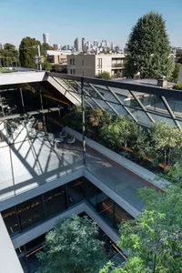 High-angle view of a modern multi-level rooftop terrace featuring a lush indoor courtyard garden, glass railings, concrete surfaces, and a panoramic city skyline in the background.