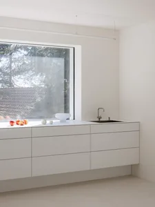Modern minimalist kitchen featuring handleless white matte lacquer cabinetry, integrated black sink with stainless steel faucet, and a large picture window overlooking trees and a tiled roof.