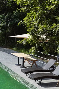 Two dark brown woven wicker lounge chairs on a grey tiled pool deck next to a swimming pool with green water, accompanied by a light wood table and bench under a beige triangular shade sail, surrounded by lush green trees.