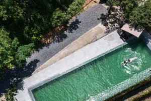 Bird's eye view of a rectangular swimming pool with emerald green tiles, light gray stone deck, gravel path, and dense tropical vegetation surrounding it, with a woman swimming.