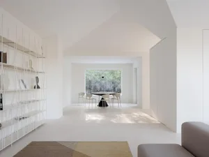 Bright minimalist white dining room featuring a black conical dining table, light wood chairs, tall white wireframe shelving unit, and a geometric brown and yellow rug in the foreground.