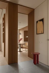 Modern minimalist entryway featuring warm walnut wood cabinetry, a glossy red cylindrical side table, and a view into a light-filled dining area with a round pedestal table and cane chair.