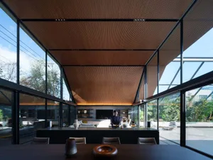 Modern dining room with dark wood table and chairs, black-framed floor-to-ceiling glass walls, and a warm wooden slatted ceiling that extends outward, with a woman standing at the counter in the background.
