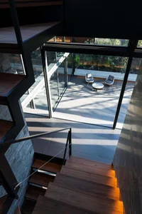 Modern walnut staircase with black metal railing descending towards a sunlit courtyard featuring two lounge chairs and a round table, viewed from above.