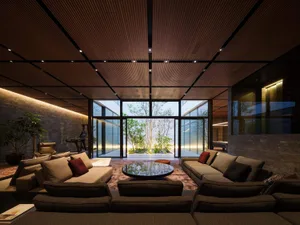 Modern luxury living room featuring dark wood slat ceiling, large sectional sofas, round coffee table, and floor-to-ceiling glass doors opening to a private garden with trees.