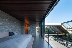 Modern minimalist bedroom with textured grey stone wall, wooden platform beds with grey linens, dark wood slat ceiling, and large glass walls opening to a bright outdoor terrace.