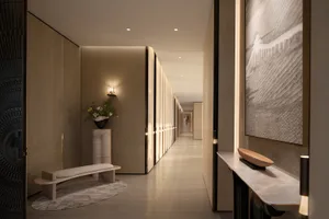 Modern minimalist luxury hallway featuring a travertine console table, wall sconces, large abstract artwork, and a curved stone bench with floral arrangement on a marble pedestal.