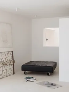 Minimalist interior corner with a black leather daybed on casters, textured white art pieces, a stack of books, and grey slippers on a woven mat.