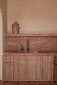 Modern minimalist kitchen featuring a terracotta marble backsplash and countertop, light wood cabinetry, double stainless steel sinks with a tall gooseneck faucet, and a terracotta urn on the shelf against a textured plaster wall.