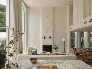 Bright double-height living room featuring a sculptural white plaster fireplace, curved travertine coffee table, white ribbed sofa, and wooden dining chairs with large arched windows overlooking greenery.
