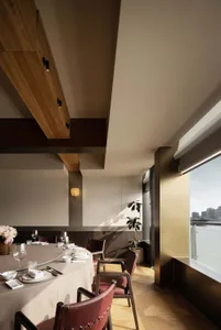 Modern elegant dining room featuring a large round table with beige tablecloth, burgundy leather chairs, walnut wood ceiling beams, and a large window with harbor view.