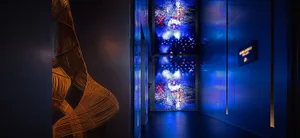 Modern exhibition corridor featuring large-scale digital art of colorful fireworks on LED screens, dramatic blue lighting, and an orange-brown woven sculptural installation on the left.