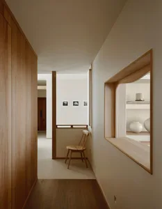 Modern minimalist hallway featuring light oak wood paneling, a built-in curved display niche with white ceramic vases, and a wooden chair against a white wall with small framed photographs.