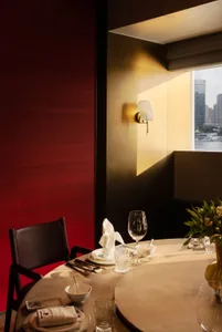 Elegant round dining table set for two with white tableware, crystal glasses, and a floral arrangement, positioned beside a rich red walnut wall and a large window overlooking a harbor with boats and city buildings.
