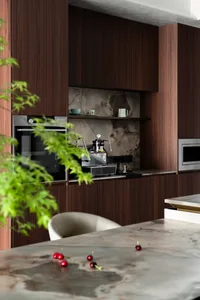 Modern luxury kitchen featuring dark walnut wood cabinetry, a built-in espresso machine on a marble countertop, and a dramatic marble backsplash with open shelving displaying coffee cups.