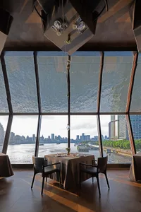 Modern fine-dining table with black leather chairs positioned in front of enormous angled windows revealing a river and dense city skyline, under a striking geometric wood-paneled ceiling feature.